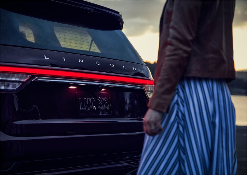 A person is shown near the rear of a 2023 Lincoln Aviator® SUV as the Lincoln Embrace illuminates the rear lights | Mike Carpino Lincoln in Pittsburg KS