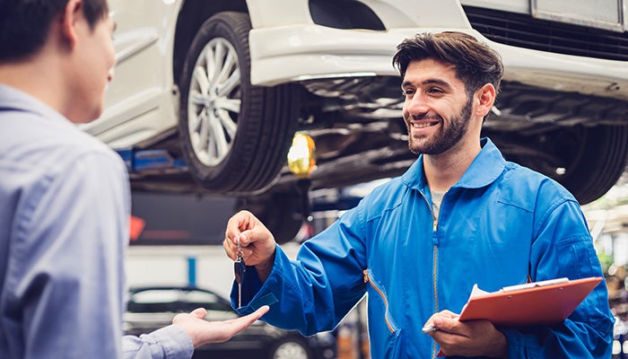 A smiling mechanic in blue overalls hands car keys to a customer.