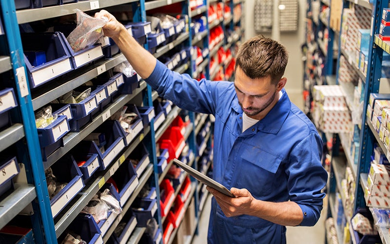 A man in a blue uniform takes an item from a bin on a warehouse shelf while looking at a tablet.