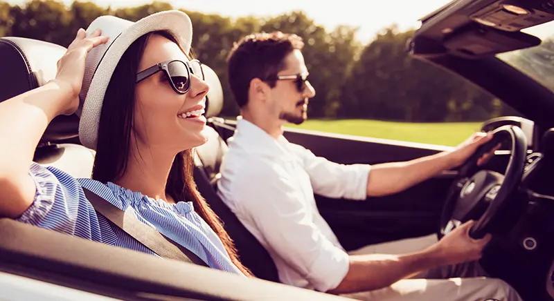 A smiling woman in a hat and sunglasses rides in a convertible while a man drives.